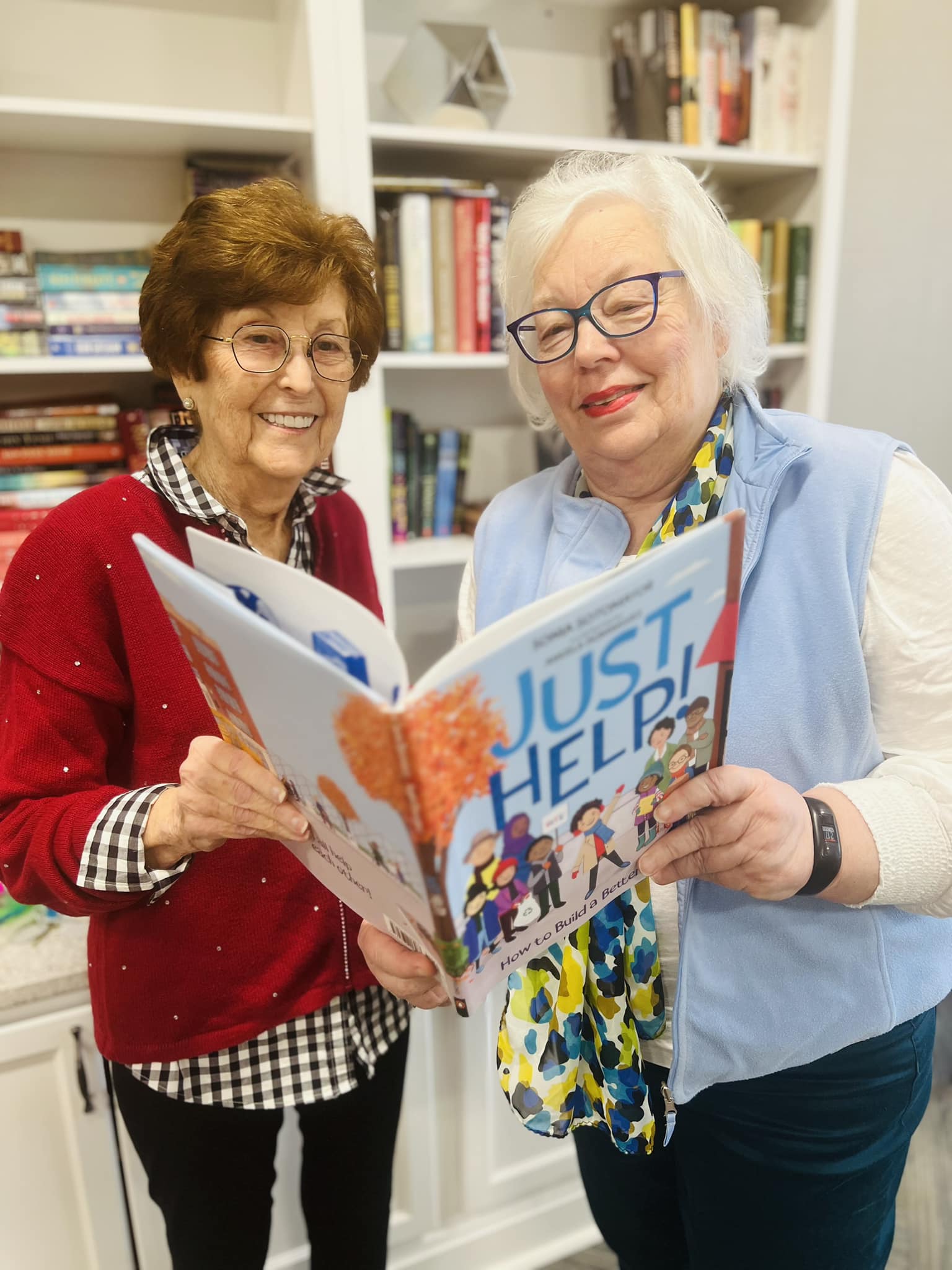 Two senior residents smiling as they read a book titled 'Just Help! How to Build a Better World' together in a cozy library setting, surrounded by shelves filled with books.