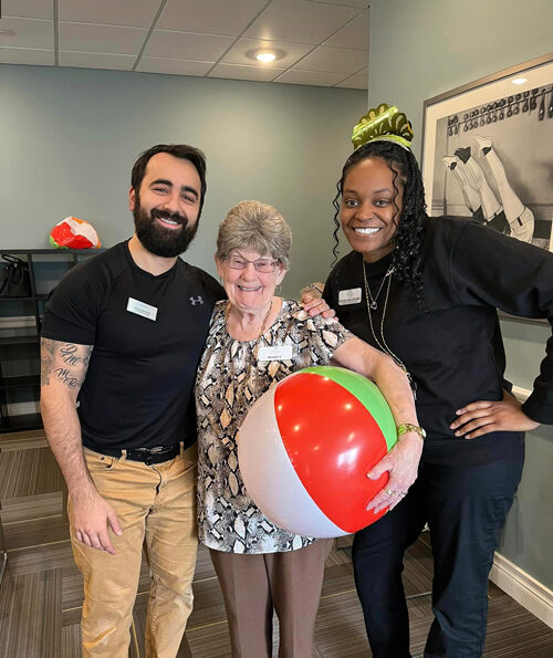 Smiling senior resident holding a beach ball stands between two staff members, one male and one female, during a festive indoor event, with all three posing happily together.