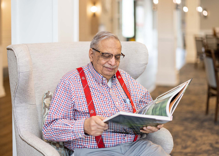 A senior resident wearing glasses and red suspenders sits in a comfortable chair, reading a large book in a senior living community, enjoying a quiet and relaxing moment.
