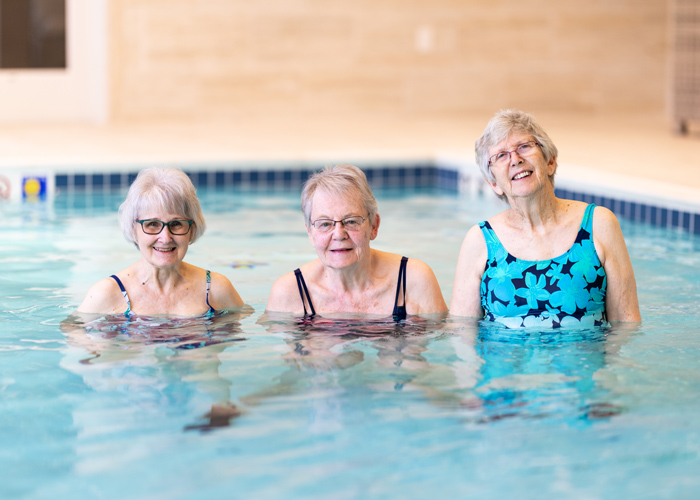 Three senior residents enjoying a swim, standing waist-deep in a clear indoor pool, smiling and wearing swimsuits, capturing a moment of relaxation and fun together.