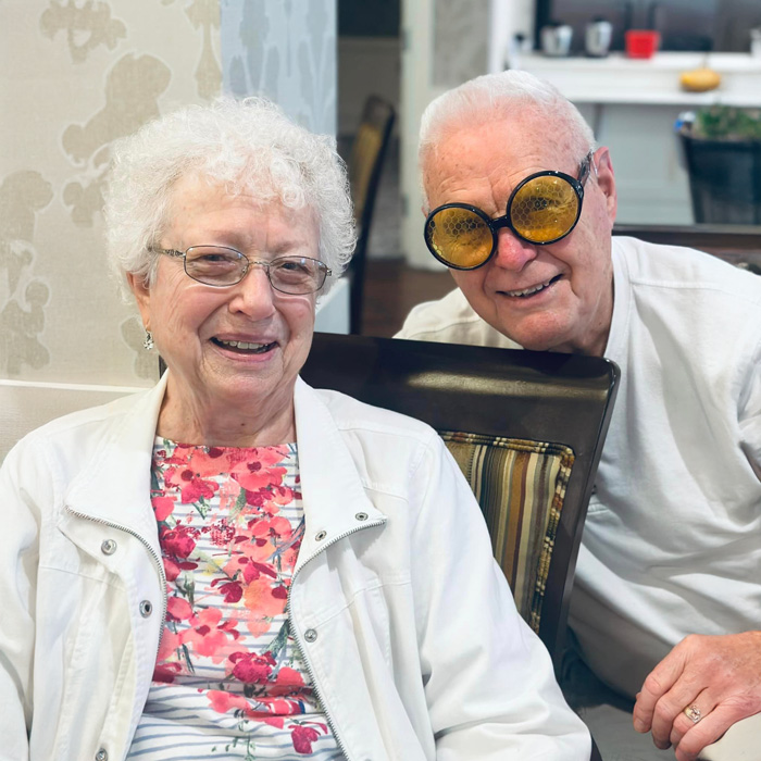 Two smiling senior residents enjoying time together, with one wearing humorous oversized yellow glasses, creating a fun and lighthearted atmosphere in the community.