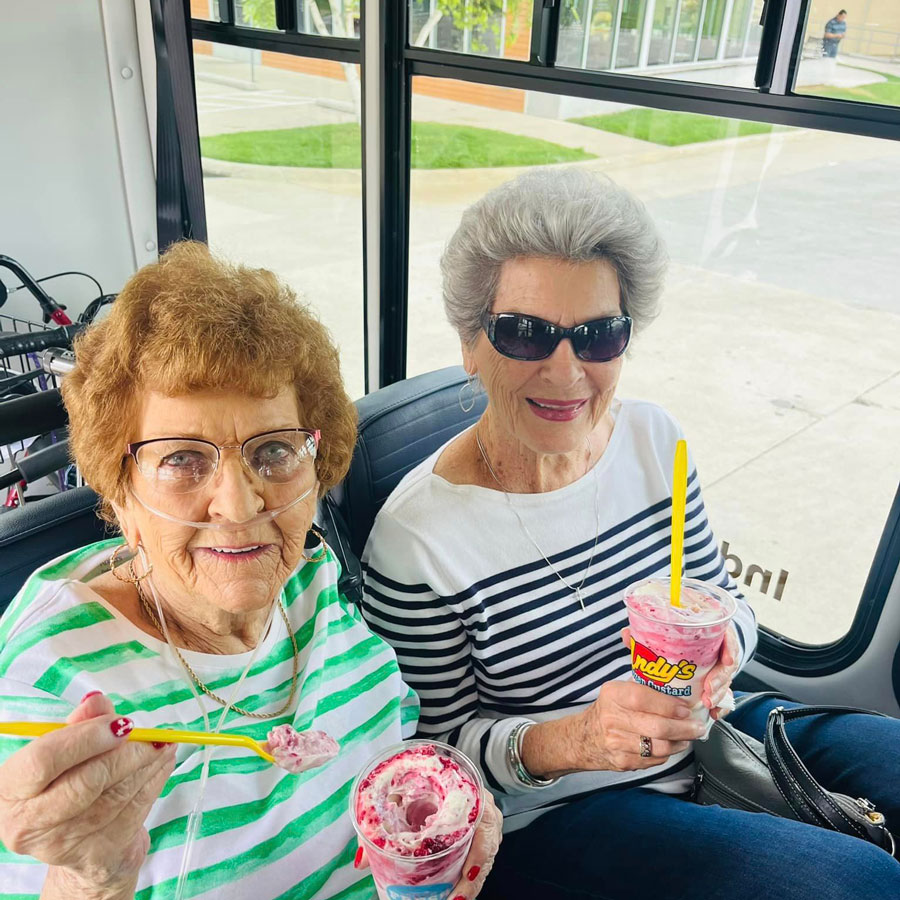 Two senior residents enjoy frozen custard treats while sitting on a bus. One wears a green striped shirt with an oxygen tube, and the other wears a black and white striped shirt.