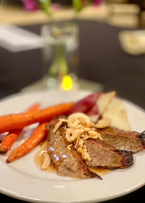 Plate of tender roasted beef topped with crispy onions, served with glazed carrots and a side of mashed potatoes, set against a dimly lit table with a candle centerpiece.