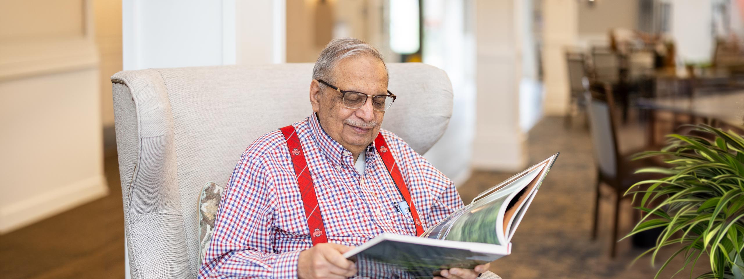 Senior resident sitting comfortably in a lounge chair, wearing red suspenders and glasses, smiling as he reads a book, representing relaxation and intellectual engagement.