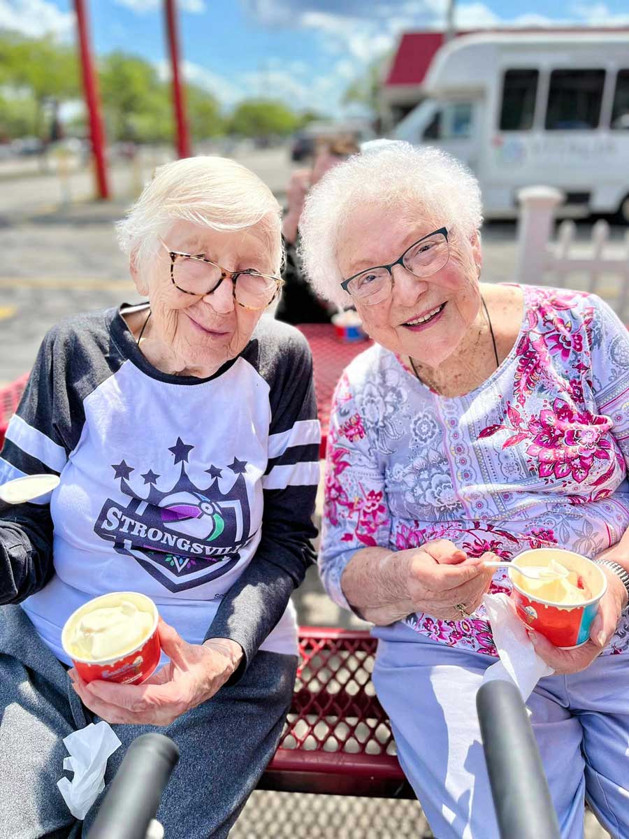 Two senior residents enjoy ice cream together at an outdoor table on a sunny day, with smiles and a Strongsville community van visible in the background.
