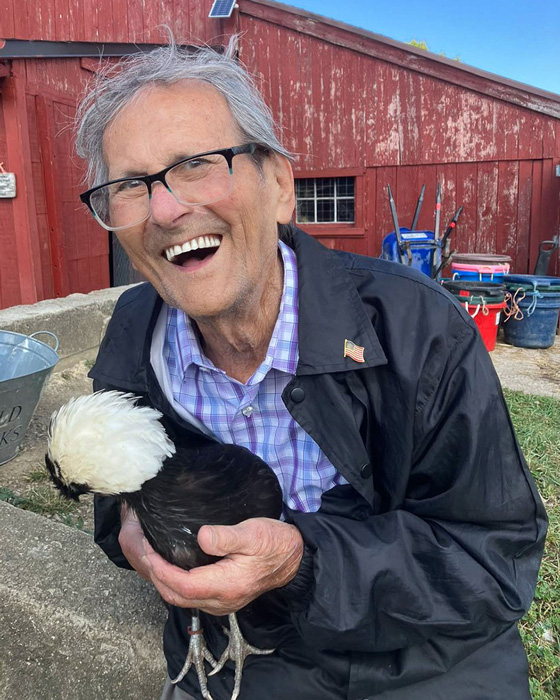 Senior resident laughing joyfully while holding a chicken during a visit to a farm, with a red barn and farming equipment in the background, enjoying an outdoor adventure.