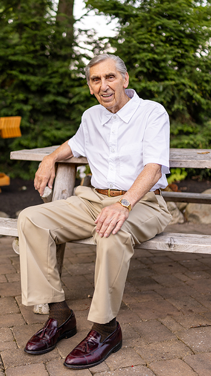 Senior resident in a white short-sleeve shirt and beige trousers, sitting on a wooden bench outdoors, smiling warmly with green trees in the background.