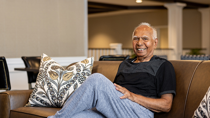 Senior resident relaxing on a brown couch, smiling in a cozy lounge area with patterned pillows, creating a welcoming and comfortable atmosphere.