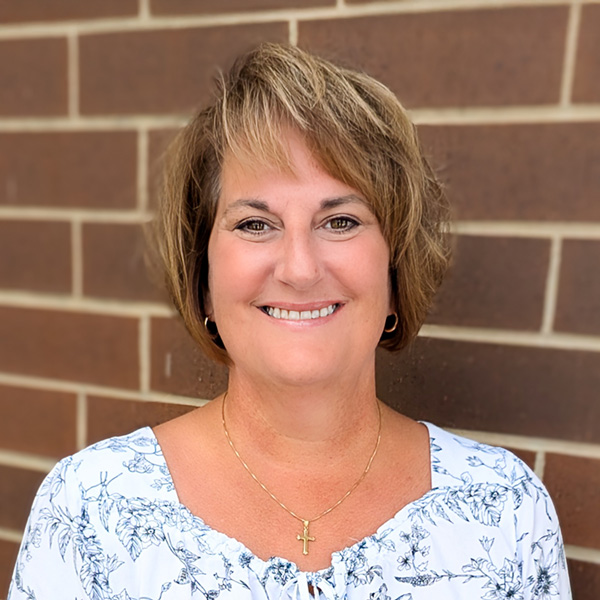 Christine Cross, Executive Director at Aura Independent Living, smiling in front of a brick wall while wearing a white floral top and gold cross necklace.