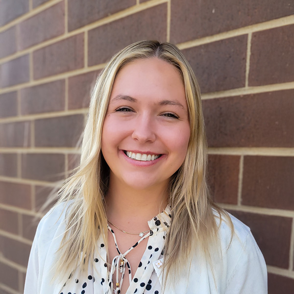 Sydney Busse, Senior Living Coordinator at Aura Independent Living, smiling while standing in front of a brown brick wall, wearing a white blouse with black polka dots and a white cardigan.