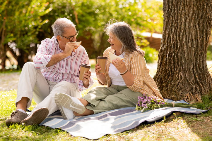 Senior couple enjoying a picnic on a blanket under a tree with coffee and sandwiches