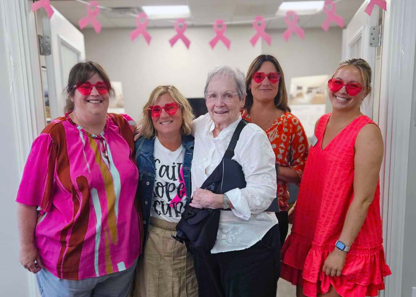 A senior woman and four team members smile in front of Breast Cancer Awareness decorations, the team members wearing heart glasses.