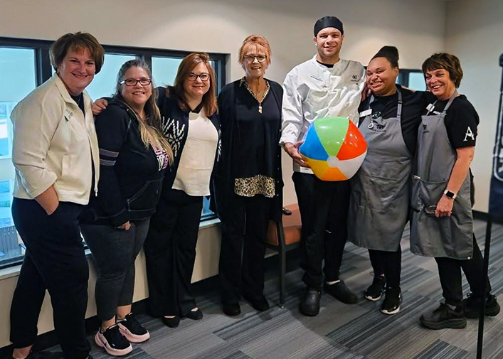 A group of team members stands together, smiling warmly. One team member in a chef coat holds a colorful beach ball as everyone gathers close for a cheerful group photo.
