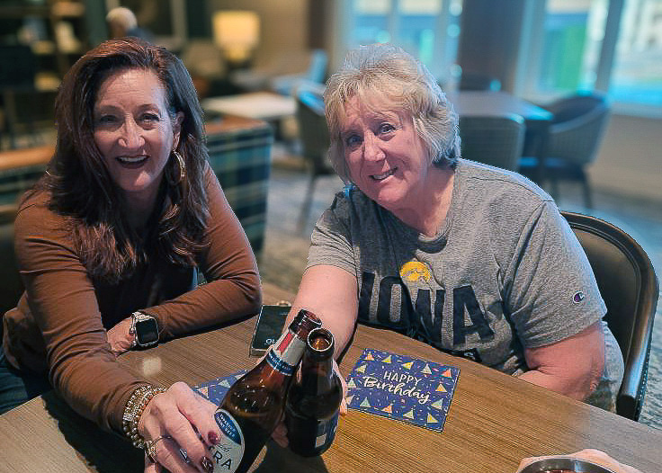 Two women sit together at a table, sharing drinks and conversation. Both smile toward the camera, creating a relaxed and friendly moment during a celebration.