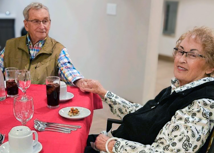A senior couple sits at a table covered with a red tablecloth, holding hands and smiling. Glasses and small plates of food are set neatly in front of them, capturing a sweet and comfortable dining moment.