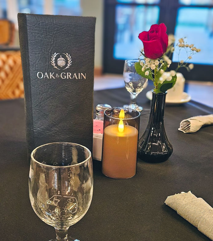 A table is set with a black tablecloth, a glass of water, a flickering candle, and a single red rose in a small vase. A menu embossed with the name Oak & Grain stands upright as soft natural light fills the background.