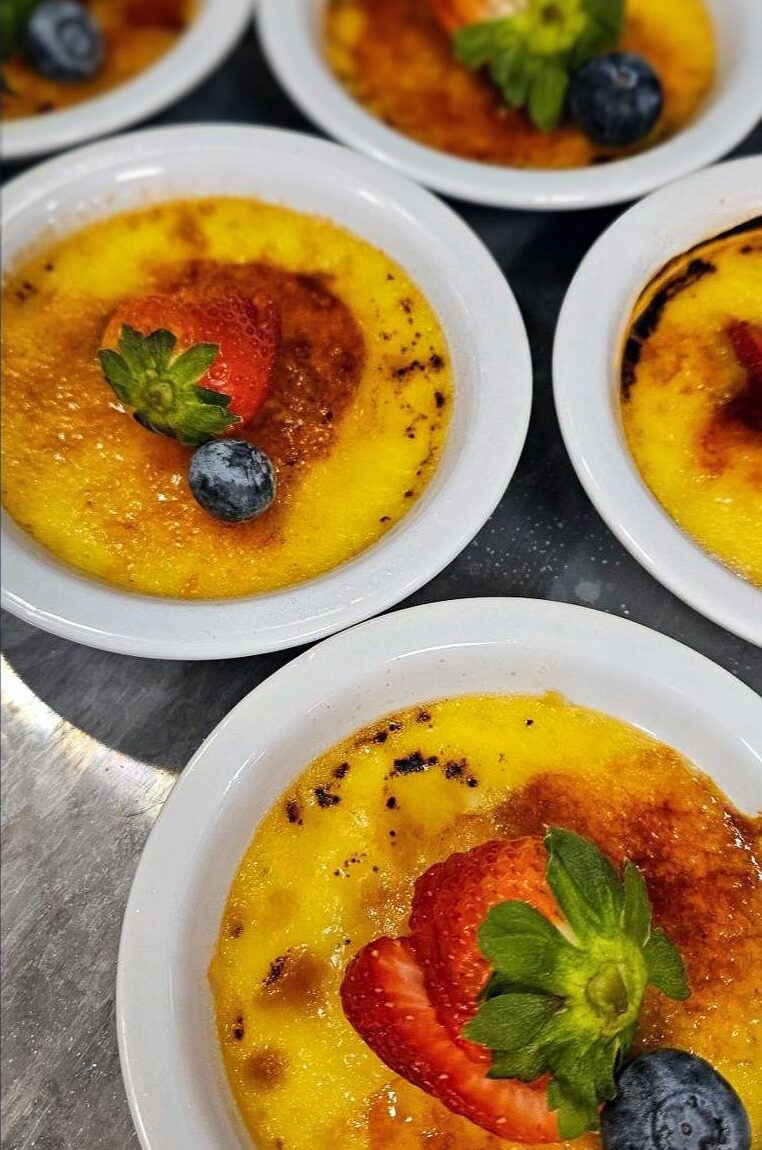 Several small bowls of crème brûlée are arranged on a tray, each topped with a fresh strawberry slice and a blueberry. The caramelized tops are golden and slightly crisp, ready to be served.