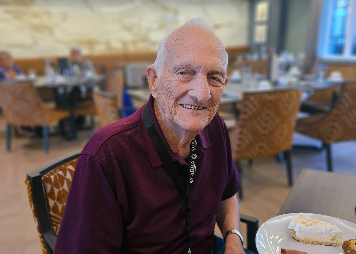 A senior resident wearing a burgundy shirt sits at a dining table, smiling gently at the camera. A slice of pie is placed on the plate in front of him as the dining room activity continues behind him.