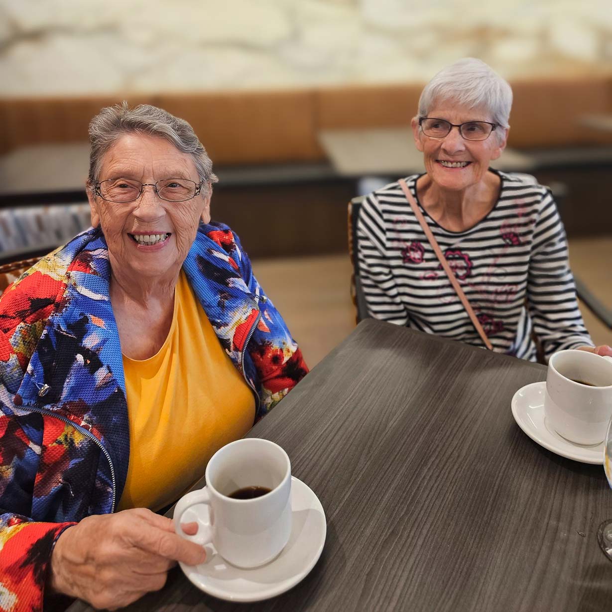 Two senior residents sit side by side at a table, each holding a warm cup of coffee. Both smile brightly, wearing colorful and patterned outfits that add to the cheerful atmosphere.