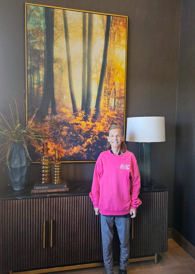A smiling woman stands in front of a large framed autumn forest painting, wearing a bright pink sweatshirt and gray pants in a warmly lit, modern room.