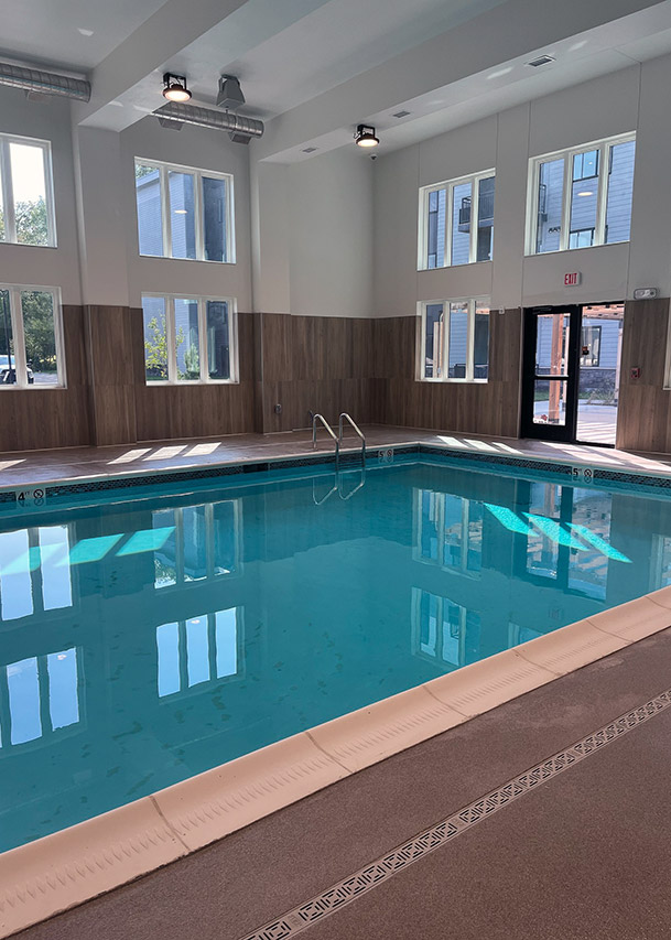 A bright indoor swimming pool with clear blue water is surrounded by tall windows, wood-accented walls, and natural light streaming into the modern, serene space.