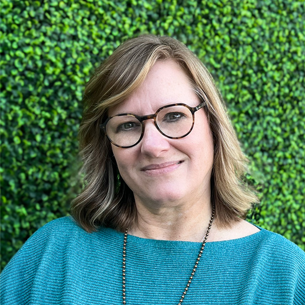 Lisa Cleland, Senior Living Director at Aura Independent Living, smiling gently in front of a vibrant green hedge wall, wearing glasses and a teal sweater.