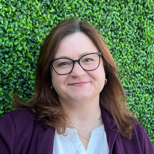 Rebecca “Becca” Cox, Senior Living Coordinator at Aura Independent Living, smiling in front of a lush green hedge wall, wearing glasses, a white blouse, and a deep purple blazer.