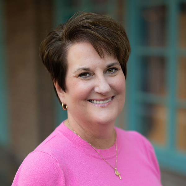 Christine Cross, Executive Director at Aura Independent Living, smiling in a professional outdoor headshot with short brown hair, a bright pink top, and softly blurred brick and teal window details in the background.