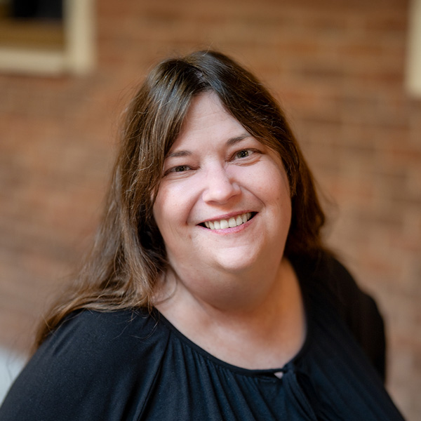 Sheri Hlavacek, Business Office Director at Aura Independent Living, smiling in a professional headshot, wearing a black top, with a softly blurred brick background.