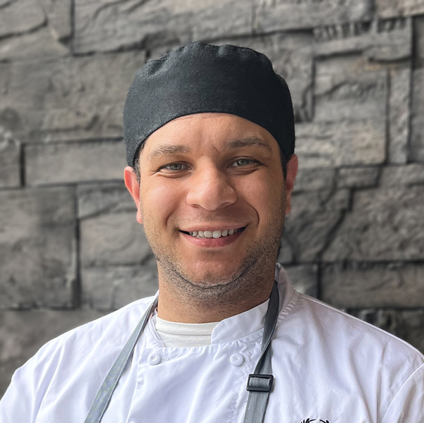DeVante Bowers, Culinary Director at Aura Independent Living, smiling in a professional headshot wearing a white chef coat and black chef cap, with a dark stone wall background.