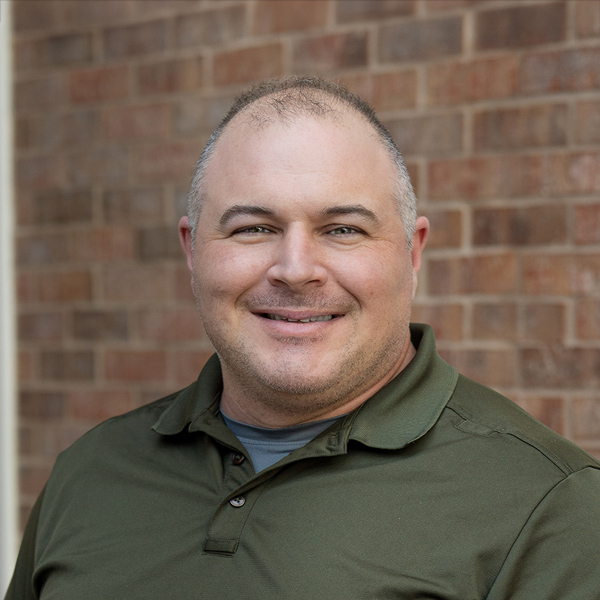 Dustin Dallenbach, Plant Operations Director at Aura Independent Living, smiling in a professional outdoor headshot with closely cropped hair, an olive green polo shirt, and a softly blurred brick background.