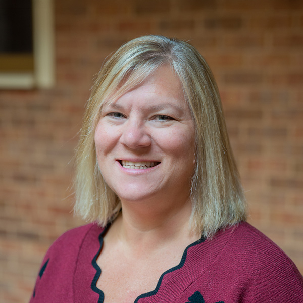 Gail McLaughlin, Resident Services Director at Aura Independent Living, smiling in a professional outdoor headshot with straight blonde hair, a burgundy sweater, and a softly blurred brick background.
