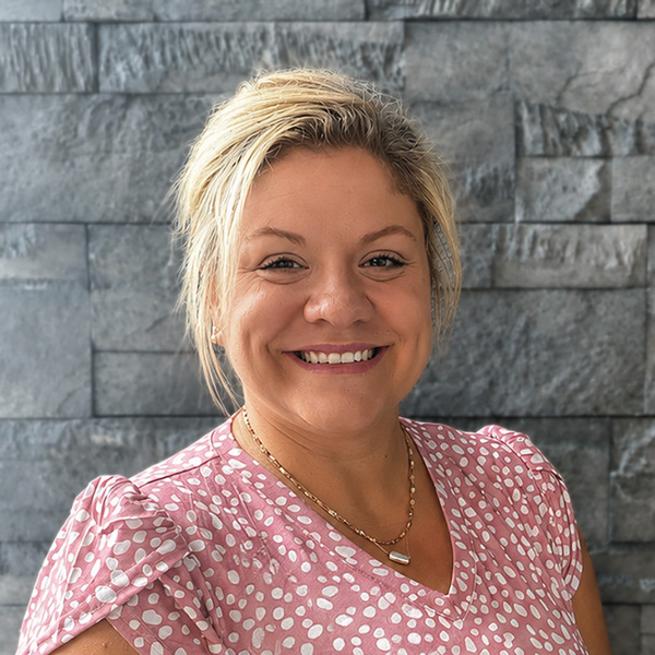 Jaymin Pirtle, Senior Living Director at Aura Independent Living, smiling in a professional headshot with short blonde hair, a pink patterned blouse, layered necklaces, and a gray stone wall background.